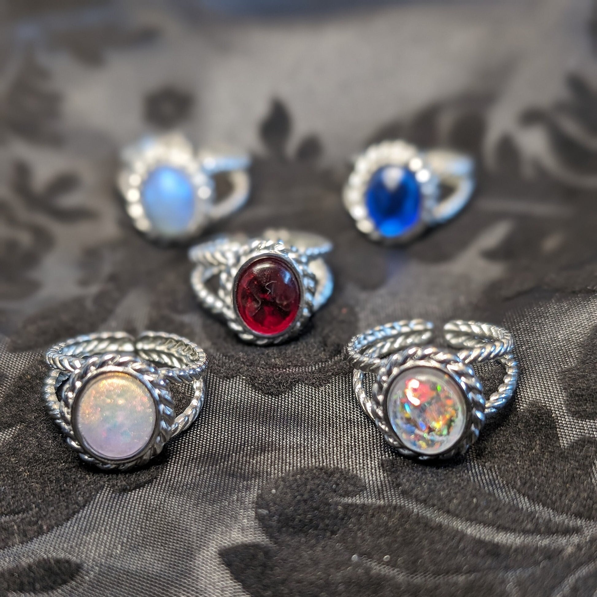 Five rings with resin cabochons and braided stainless steel bezels sit on a black fabric background