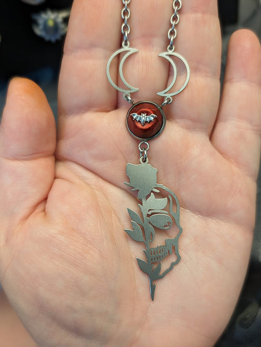 rose and half skull stainless steel charm hangs from a round, red resin and stainless steel connector, which dangles from two stainless steel crescent moon charms on a stainless steel chain. The necklace sits against the palm of a hand for scale.